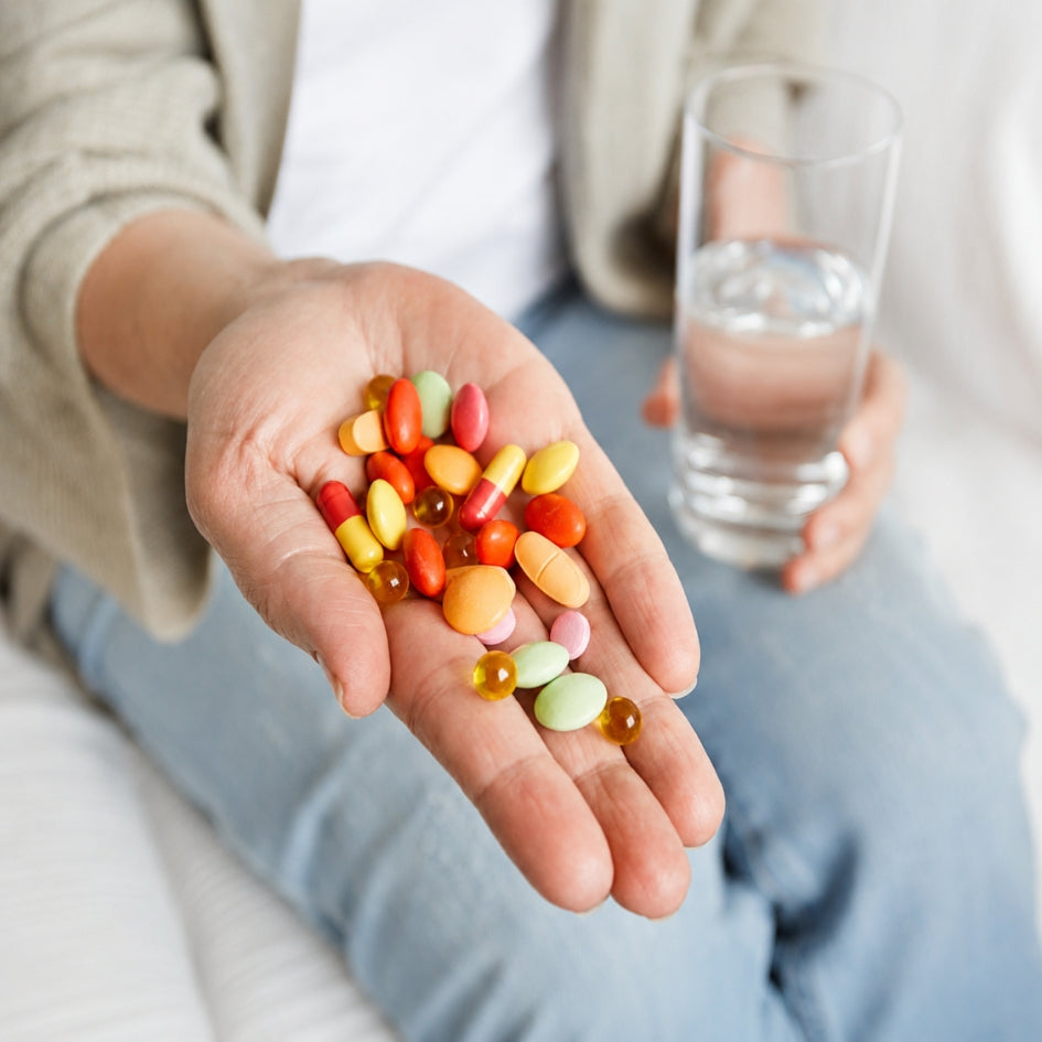 A close-up of a person's open palm holding a mix of colorful softgels and capsules, with a glass of water held in the other hand, set against a soft, neutral background.