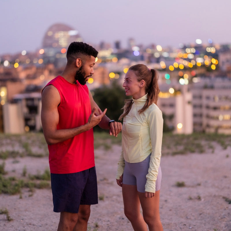 A man and a woman in athletic wear standing outdoors at dusk with a blurred city skyline in the background. The man wears a bright red sleeveless shirt and dark shorts, while the woman wears a pale yellow long-sleeve top and light gray shorts.