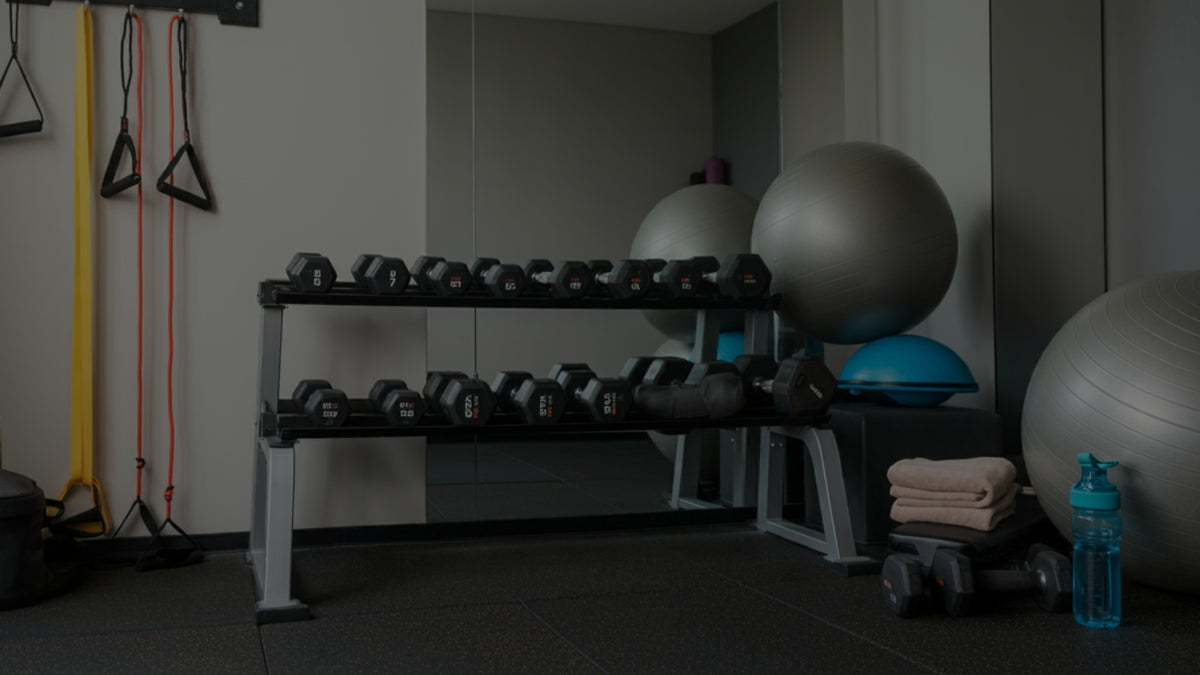 A dark gym interior with a black dumbbell rack holding multiple dumbbells, large exercise stability balls, resistance bands hanging on the left, folded towels, and a water bottle on the floor.