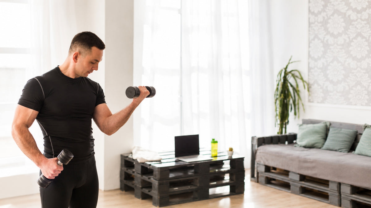 A man in a black athletic shirt and leggings holds a dumbbell in a bright living room with a sofa, pallet coffee table, and large windows.