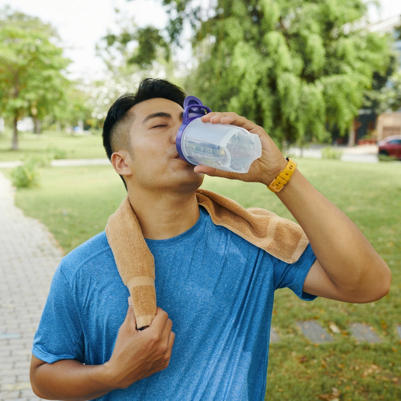 A man in a blue athletic shirt drinking from a clear plastic shaker bottle with a purple cap, with a tan towel over his shoulder, outdoors on a paved path with green trees in the background.