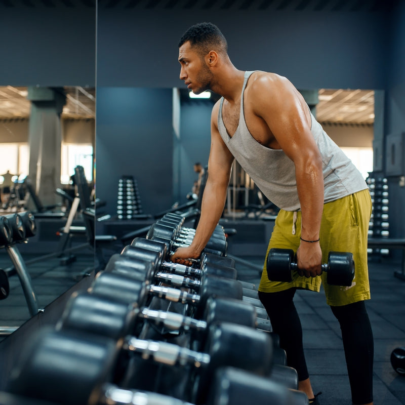 A man in a gym wearing a gray tank top, yellow shorts, and black leggings picks up a dumbbell from a rack while looking to the side.