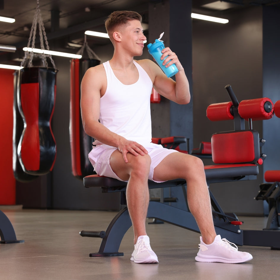 A man in a white tank top and shorts sits on a gym bench drinking from a blue water bottle, with punching bags and exercise equipment in the background.