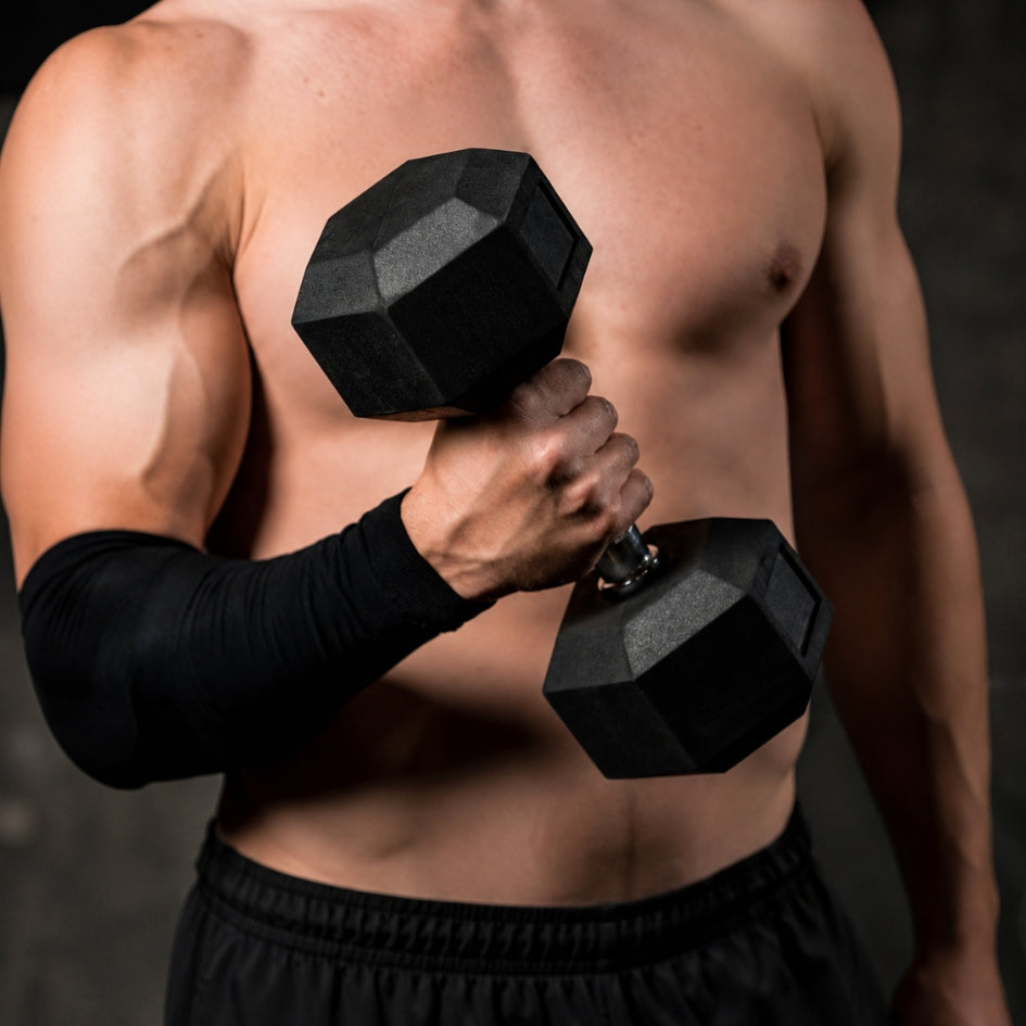A close-up of a shirtless, muscular man lifting a black hexagonal dumbbell with a black elbow sleeve on one arm against a dark studio background.