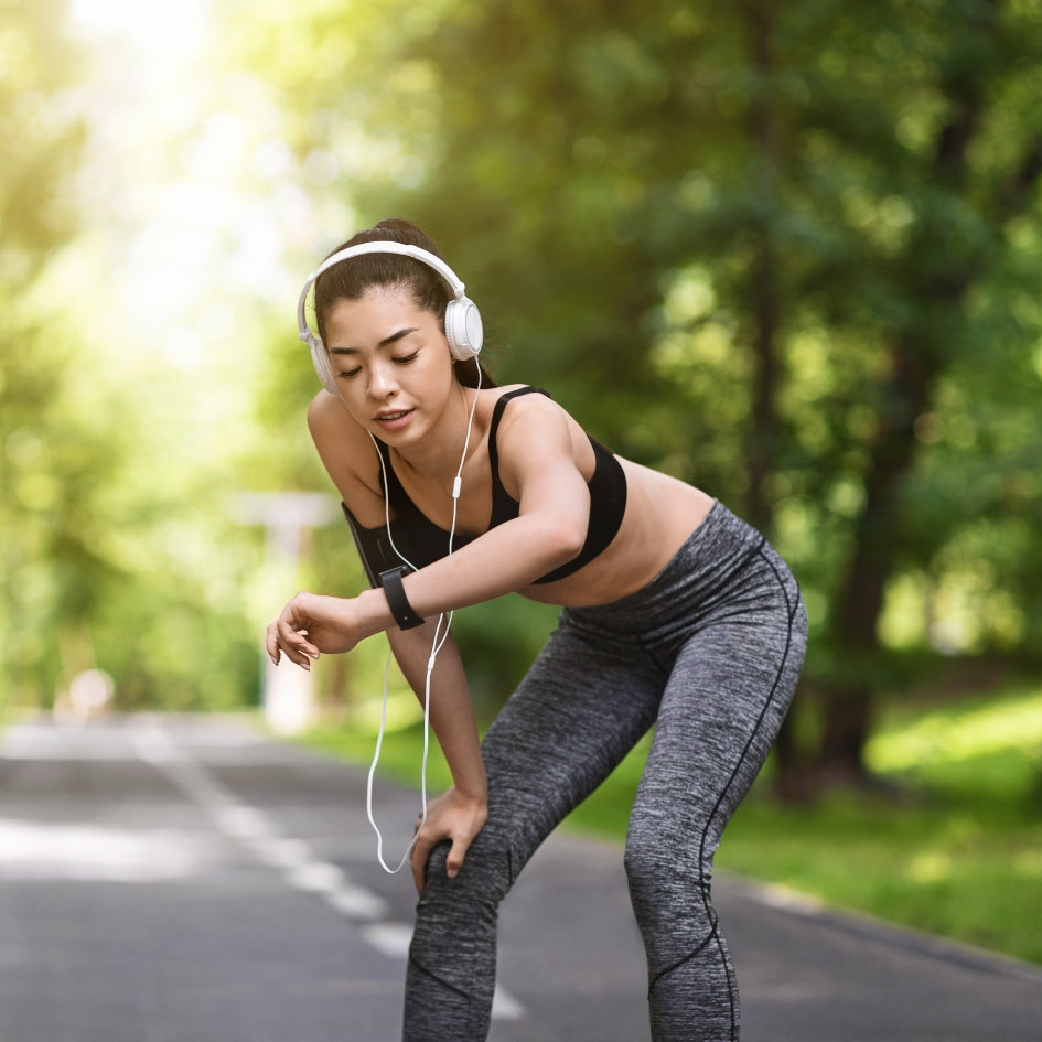 A woman in athletic wear, including a black sports bra and gray heather leggings, wearing white headphones and a smartwatch, bends over on a paved path in a sunlit park.