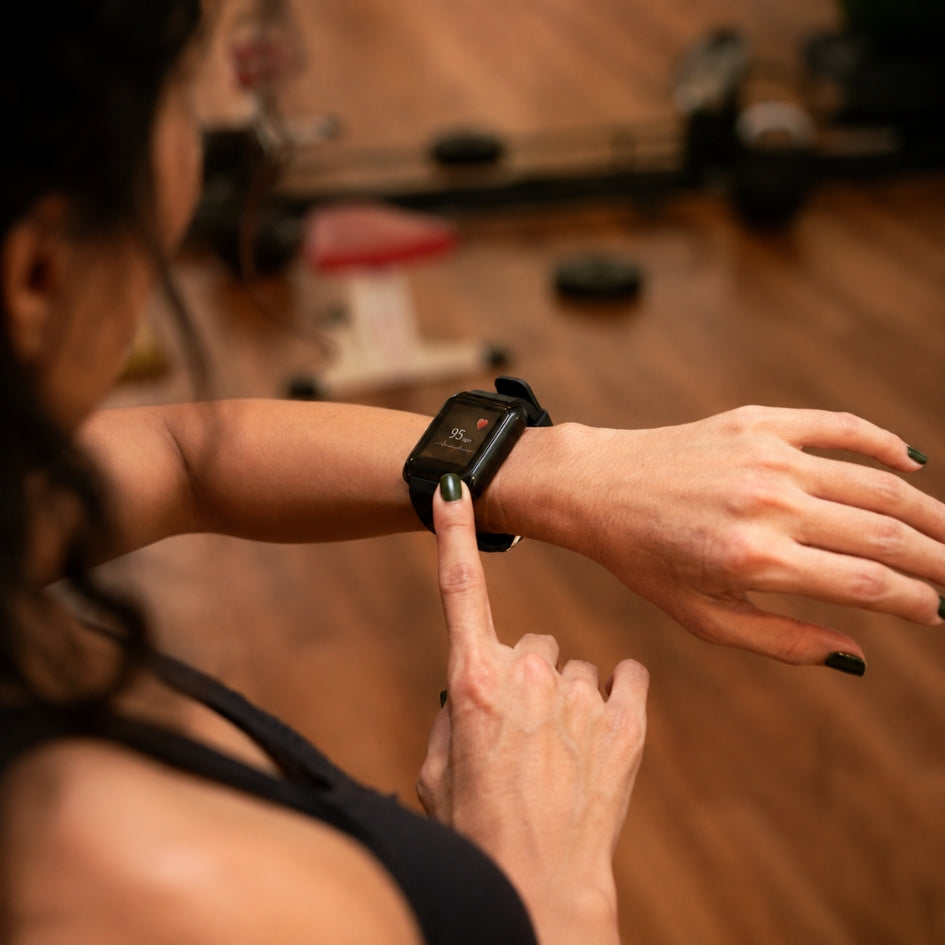 A close-up lifestyle shot of a woman in a gym setting, looking down at a black smartwatch on her wrist while pressing the screen with her finger.