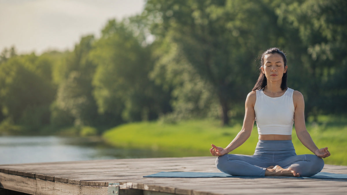 A woman meditating in a white tank top and gray leggings on a blue yoga mat, sitting cross-legged on a wooden dock with a green forest and lake in the background.