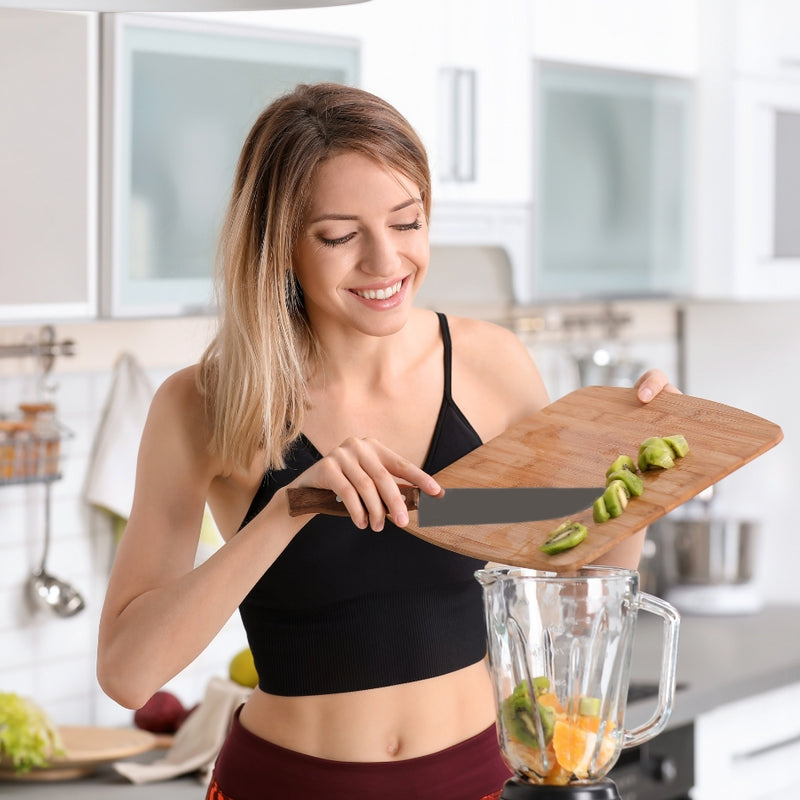 A woman in a black sports bra and burgundy leggings smiles while sliding avocado pieces from a wooden cutting board into a clear blender jar on a kitchen counter.