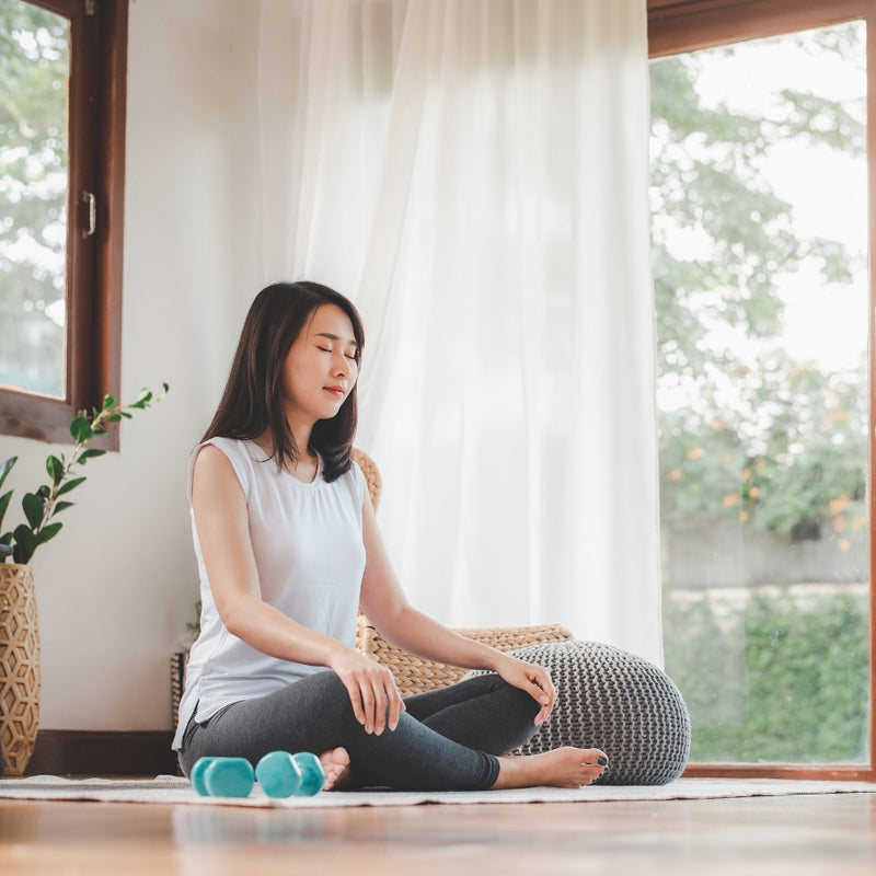 A woman sits cross-legged on a yoga mat in a bright room by large windows, wearing a white sleeveless top and dark leggings. A gray pouf cushion, small teal dumbbells, and a potted plant are visible nearby.